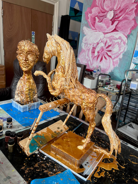 Side view of Stephanie Fuller’s gold-coloured rearing horse sculpture in progress, with a flowing mane and tail, beside a sculpted bust.