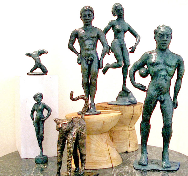 A collection of classical-style bronze sculptures depicting nude human figures and a leopard, displayed on wooden pedestals and a marble surface against a plain white background.