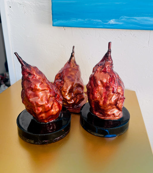 Three resin pear sculptures by Stephanie Fuller, each with a distinct textured surface in red, copper and gold tones and a long, slender stem.
